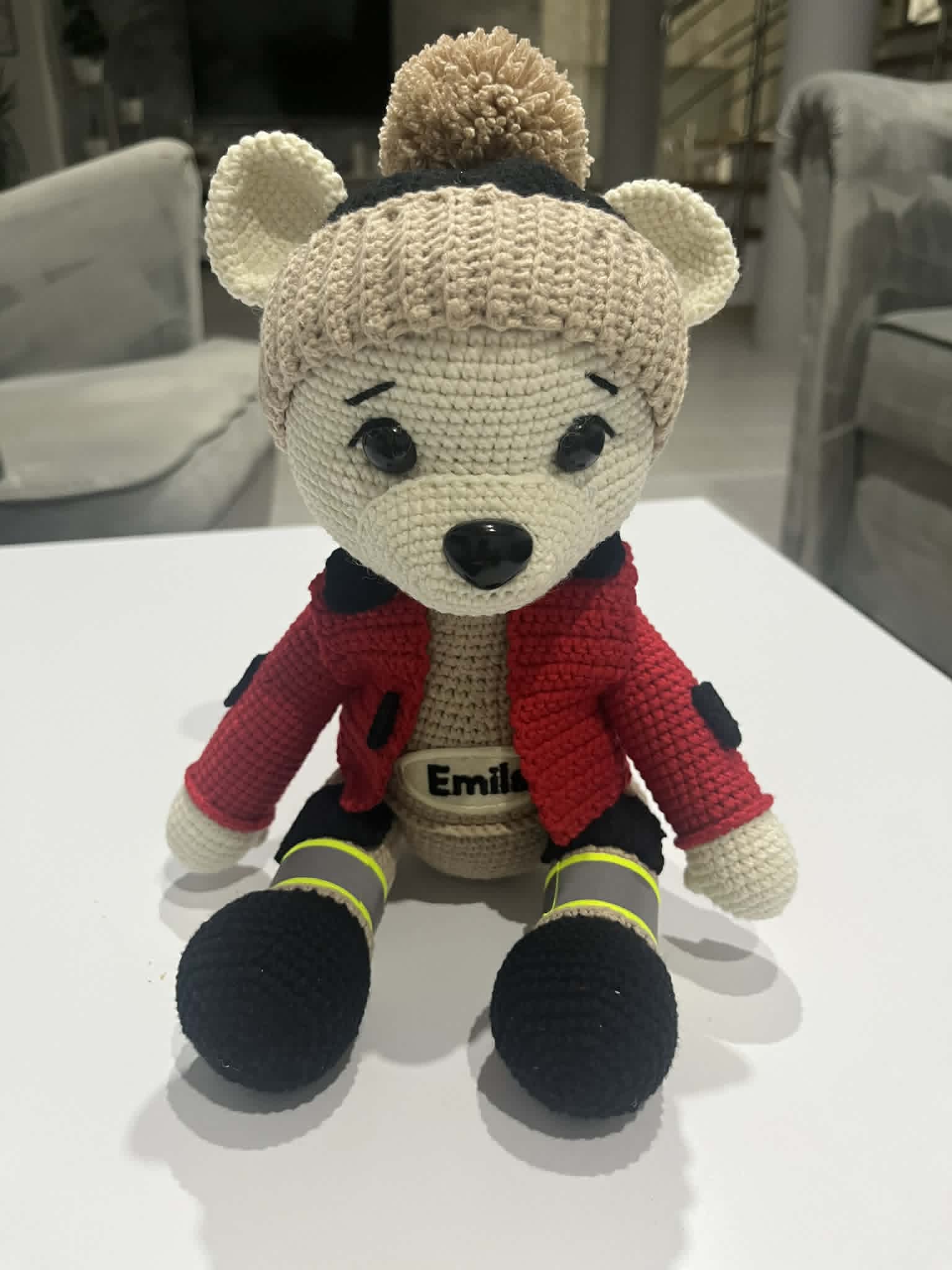 Szydełkowany miś w stroju strażaka z odblaskowymi pasami i napisem "Emils" na bluzie, wykonany ręcznie.