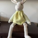 Ręcznie szydełkowana maskotka króliczka amigurumi w żółtej sukience z ozdobnym kwiatuszkiem przy uchu, siedząca na białej komodzie.