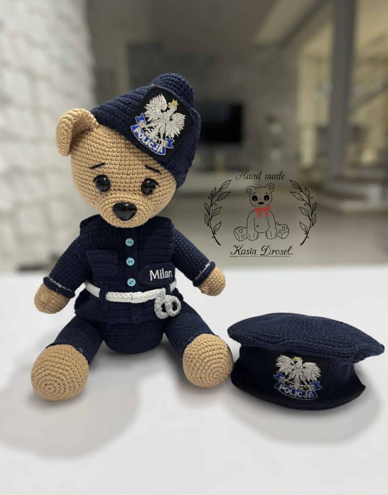 Ręcznie robiony miś amigurumi w mundurze polskiego policjanta z imieniem Milan, siedzący obok czapki policyjnej.