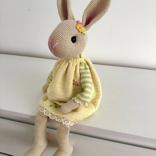 Ręcznie szydełkowana maskotka króliczka amigurumi w żółtej sukience z ozdobnym kwiatuszkiem przy uchu, siedząca na białej komodzie.