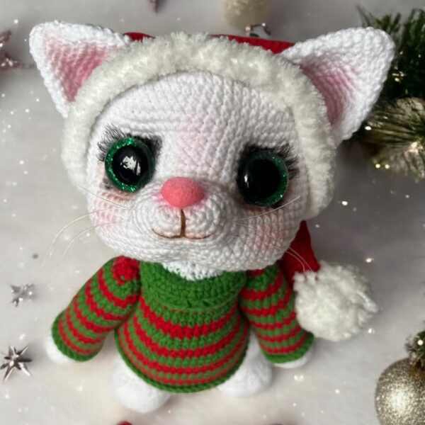 Ręcznie szydełkowana maskotka amigurumi przedstawiająca białego kotka w świątecznej czapce Mikołaja i zielono-czerwonym sweterku, obok znajduje się szydełkowany lizak w kształcie laski.