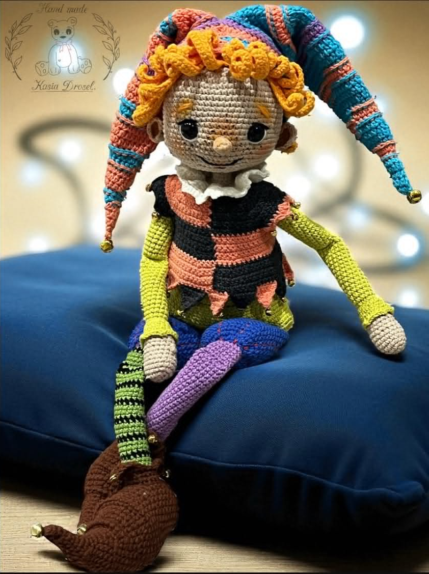 Ręcznie szydełkowana lalka amigurumi przedstawiająca kolorowego błazna w wielobarwnym stroju i czapce z dzwoneczkami, siedząca na niebieskiej poduszce.