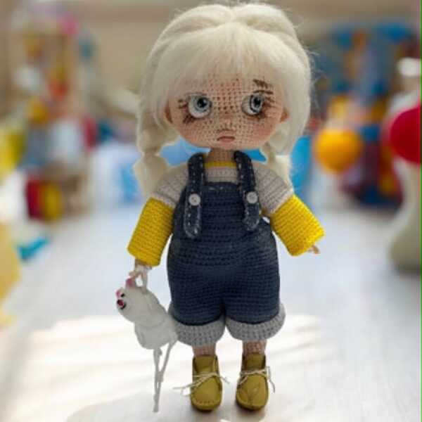 Ręcznie szydełkowana lalka amigurumi z jasnymi włosami, ubrana w żółtą bluzkę i jeansowe ogrodniczki, trzymająca małą białą maskotkę.