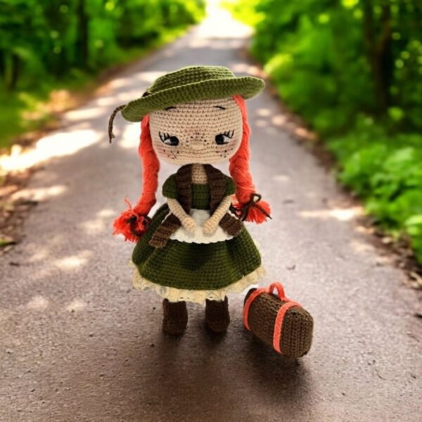 Ręcznie szydełkowana lalka amigurumi z rudymi włosami w warkoczach, ubrana w zieloną sukienkę, kapelusz i kamizelkę, z małą walizką obok, stojąca na leśnej ścieżce.